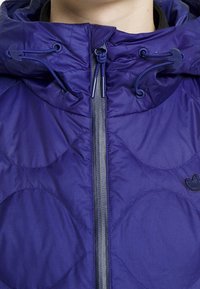 Täckvävd lila jacka med hög krage, framdragkedja och dragsko i huvan. Har cirkulära mönster och en liten broderad logotyp.