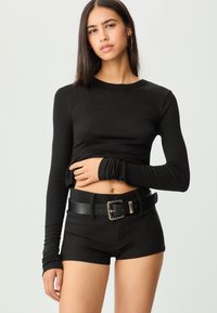 Schwarz geripptes Langarmoberteil, kombiniert mit hochgeschnittenen schwarzen Shorts und einem breiten schwarzen Gürtel mit einer silbernen Schnalle. Sanfte Stofftextur.