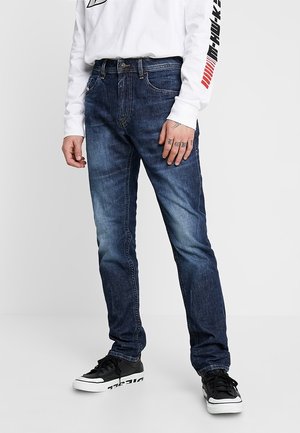 Osoba ubrana w ciemnoniebieskie spodnie jeansowe z wyblakłym efektem, czarno-białe trampki oraz białą koszulę z długim rękawem z czarnym i czerwonym napisem na rękawie.