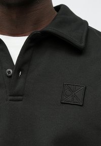 Ciemnozielona koszulka polo z teksturowanej tkaniny, z płaskim kołnierzykiem i pojedynczym zapięciem na guzik. Haftowane logo na piersi.