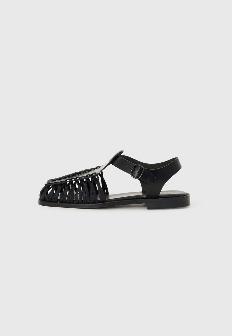 Sandal in pelle nera con design aperto, dettagli in frange sulla punta e una fascia regolabile sul tallone con suola piatta.