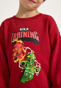 Sweat-shirt rouge mettant en avant un graphisme vibrant de deux ninjas en action, l'un rose et l'autre vert, avec des accents de flammes et d'éclairs.