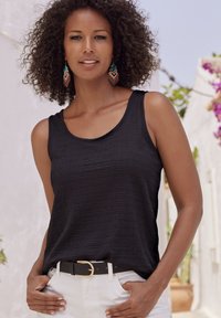 Zwarte gestructureerde tanktop met brede bandjes, gecombineerd met witte broek en een zwarte riem met een gouden gesp. Accessoires omvatten kleurrijke oorbellen.