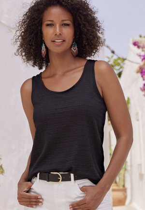 Débardeur noir texturé avec de larges bretelles, associé à un pantalon blanc et une ceinture noire avec une boucle dorée. Les accessoires comprennent des boucles d'oreilles colorées.