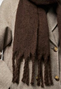 Brun uldnål med en blød, tekstureret overflade, frynset kanter og en moderat bredde, draperet over en lys grå frakke med knapper.
