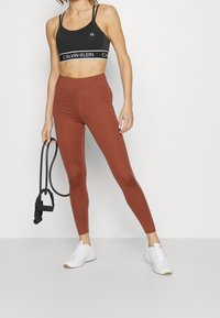Top de sport noir avec logo Calvin Klein, associé à un legging taille haute de couleur rouille. Le modèle tient une bande de résistance noire, portant des baskets blanches.