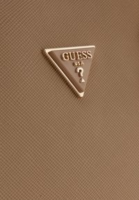 Superficie testurizzata beige con un emblema triangolare dorato contenente la scritta "GUESS USA" e un design a forma di punto interrogativo al centro.