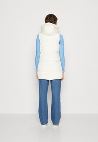 Puffy cream vest with a hood, featuring horizontal stitching, worn over a light blue ribbed long-sleeve shirt and paired with blue jeans.