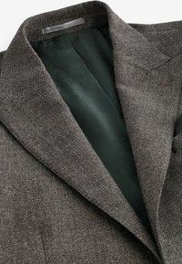 Smeđi blazer s uzorkom sa teksturiranim vanjskim slojem i glatkom tamnozelenom podstavom. Istaknute karakteristike uključuju krojeni ovratnik i suptilne detalje šivanja.