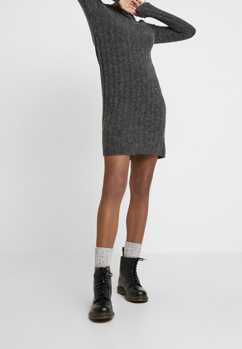 Abito maglione grigio a coste con maniche lunghe, spacco laterale, abbinato a stivali neri con lacci e calzini di lana beige.