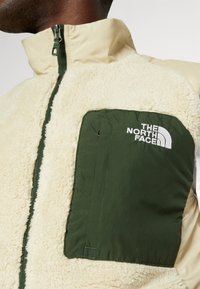 Uomo che indossa una giacca in sherpa fleece color crema con tasca a cerniera di colore verde scuro sul petto e il logo The North Face sulla tasca.