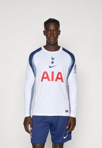 Langærmet hvid trøje med blå detaljer, rød "AIA" logo, Tottenham emblem, kombineret med marineblå shorts med en hvid Nike swoosh.