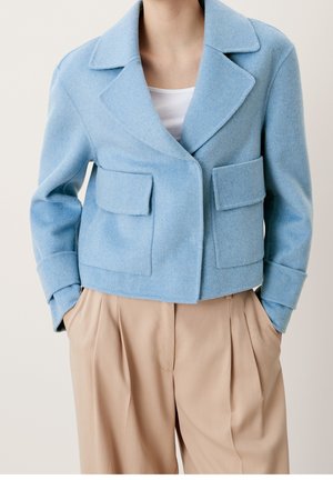Grijsblauwe wollen korte blazer met een geknipt revers, twee voorste opgestikte zakken en gestructureerde schouders. Gecombineerd met beige geplooide broek.
