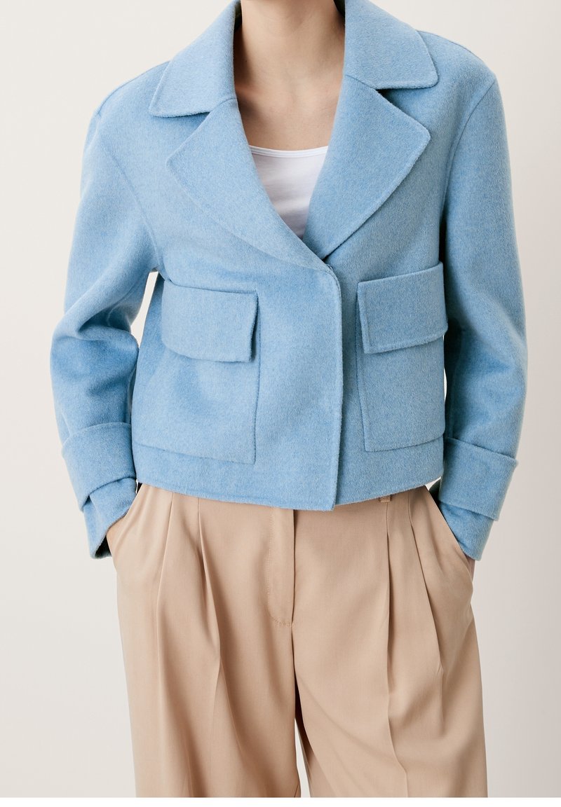 Kortad blå ullblazer med slagkrage, två framsida fackfickor och strukturerade axlar. I kombination med beiga veckade byxor.