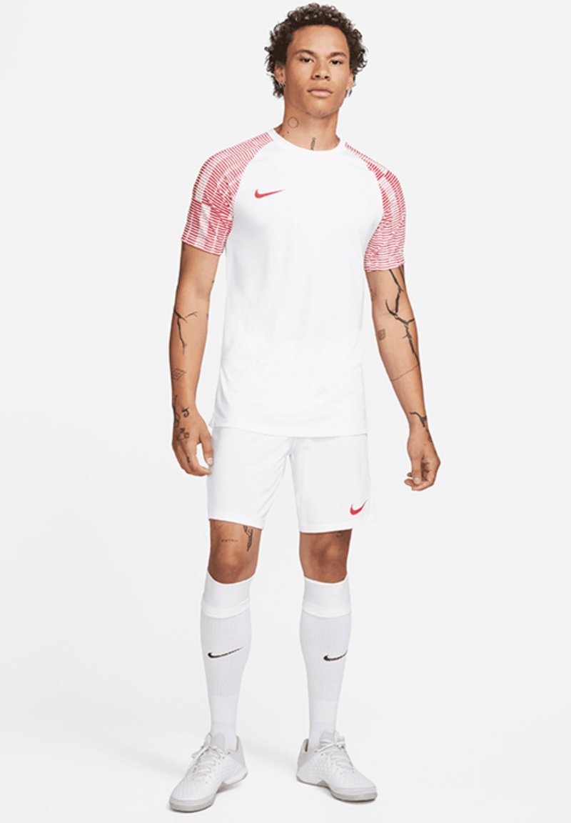 Wit voetbalshirt met korte mouwen en rode gestreepte accenten, gecombineerd met witte shorts en kniekousen. Bevat het Nike-logo in rood en zwart.