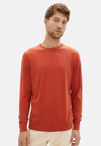 Suéter de punto rojo con cuello redondo, mangas largas, puños y dobladillo acanalados. Textura suave y minimalista con un logo en el pecho.