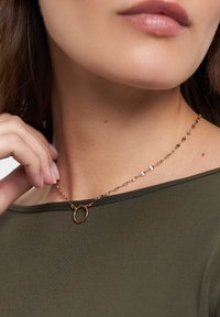 Rosaguld halskæde med en rund vedhæng, kædedesign med løse led, glat overflade, båret over en mørkegrøn top.