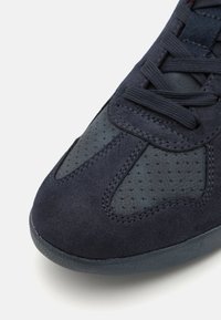 Chaussure en daim marine avec un panneau en maille sombre texturé, comprenant un bout rond, des lacets et des détails de couture contrastants. Semelle en caoutchouc lisse.