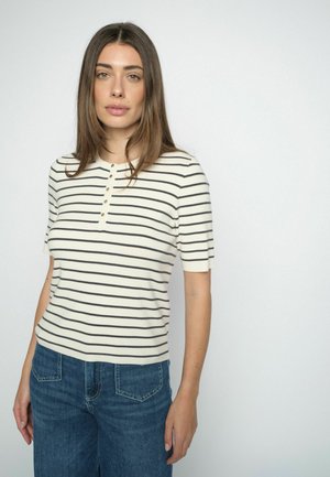 Woman with straight brown hair wearing a short-sleeve white top with black horizontal stripes and blue jeans, standing against a plain light background.