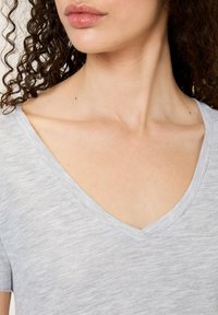 Pilka V formos kaklo marškinėliai su minkštu melange tekstūru. Yra trumpomis rankovėmis ir laisvu siluetu, išryškinančiu kaklo kaulą ir apykaklės liniją.