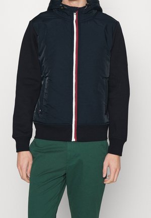 Homme portant une veste à capuche bleu marine et noire avec une fermeture éclair rouge et blanche, associée à un pantalon vert sur un fond uni.