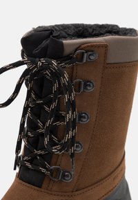 Bota de invierno de gamuza marrón con un collar acolchado negro, cordones a rayas negros y beige, ojales de metal y una suela de goma texturizada.