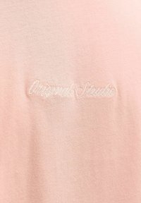 Kerge roosa puuvillane kangas, millel on broderitud tekst "Original Studio" veidi tumedamas toonis. Tekstuur on sile ja pehme.