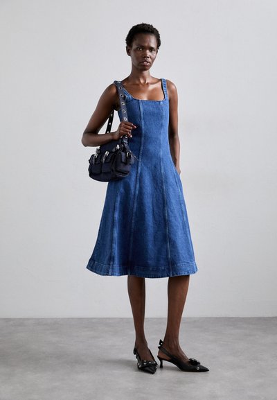 Denim jurk met een vierkante halslijn, aansluitend lijfje en uitlopende rok. Model houdt een kleine donkere handtas met meerdere zakken vast. Zwarte hakken.