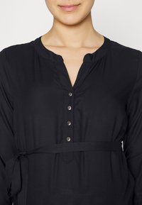 Vestido negro de botones con cuello, que cuenta con cinturón atado a la cintura, detalles de botones plateados y una textura de tela lisa.