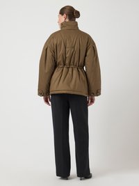 Jachetă din material moale de culoare maro, cu guler înalt, talie strânsă și manșete cu nasturi. Asortată cu pantaloni largi de culoare închisă și tocuri elegante.