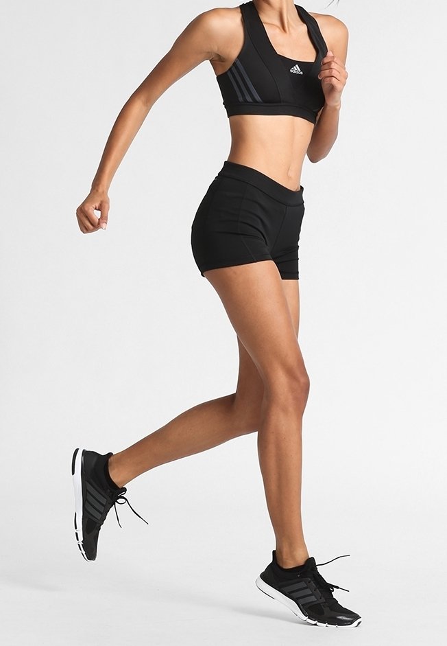 Femme courant portant un soutien-gorge de sport Adidas noir, un short noir et des chaussures de course noires sur un fond clair uni.