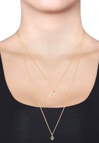 Guldpläterade halsband med en fågelberlock och en fredssymbolberlock, med fina kedjor och en slät yta, bärs på ett mörkt plagg.