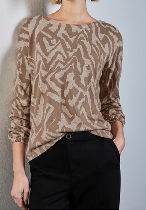 Femme portant un haut à manches longues beige et brun clair à motifs et un pantalon noir, debout contre un mur de couleur claire.