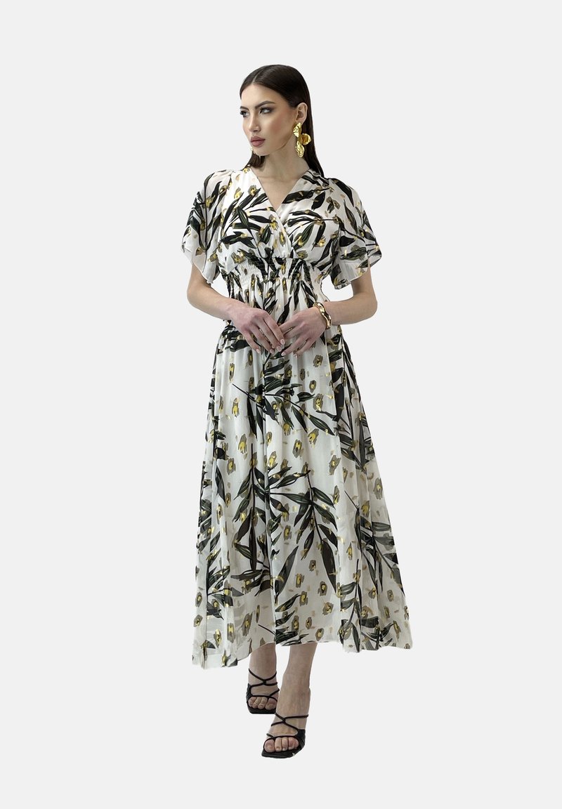 Robe maxi blanche à motifs de feuilles noires et jaunes, à manches courtes, taille smockée et tissu fluide. Associée à des sandales noires à lanières.