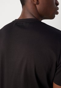 Zwarte katoenen T-shirt met een gladde textuur, ronde halslijn en subtiele logodetail op de achterkant, gepresenteerd tegen een effen achtergrond.