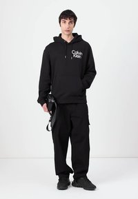 Black hooded sweatshirt with front pocket, featuring a white Calvin Klein logo. Paired with black cargo pants and black sneakers.