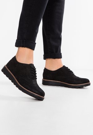 Personne portant des chaussures à lacets noires texturées et un pantalon noir retroussé, debout sur un sol blanc.
