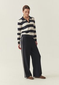 Pull rayé bleu marine et blanc avec col, associé à un pantalon large noir avec des bandes blanches sur les côtés et des chaussures marron.