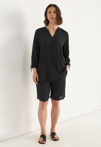 Kvinde stående mod hvid væg iført sort langærmet tunika med blonde detaljer og matchende knælange shorts samt sorte klip-klapper.