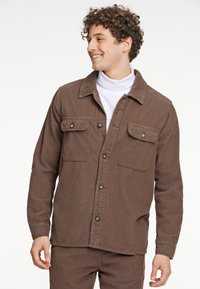 Chaqueta de camisa de corduroy marrón con frente de botones, dos bolsillos en el pecho y una superficie texturizada. Modelo lleva un cuello alto blanco debajo.