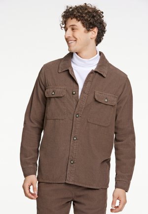 Chaqueta de camisa de corduroy marrón con frente de botones, dos bolsillos en el pecho y una superficie texturizada. Modelo lleva un cuello alto blanco debajo.