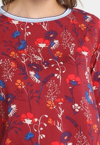 Top a fiori rossi con scollo rotondo e bordino azzurro chiaro. Il design include diversi fiori in tonalità di rosso, blu e bianco.