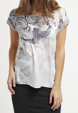 Frau trägt eine kurzärmelige Bluse mit grauen und dunklen Blumenmustern, kombiniert mit einem schwarzen Rock vor einem schlichten Hintergrund.