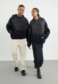 Deux mannequins portent des sweats à capuche avec un design en dégradé noir. L'homme les associe à un pantalon beige, tandis que la femme porte une jupe noire et des bottes.