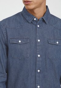 Mørk denimskjorte med spredt krage, to brystlommer og hvite knapper; strukturert stoff med en subtil falmet finish.