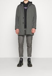 Persona in piedi contro uno sfondo bianco, indossante un cappotto grigio, una felpa nera con design viola, pantaloni grigi a maniche risvoltate, calzini bianchi e stivali di pelle neri.