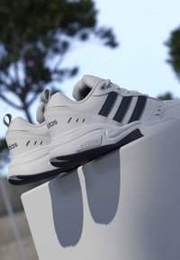Paire de baskets blanches Adidas avec des rayures noires reposant sur une surface claire en plein air sous un ciel dégagé avec des arbres flous en arrière-plan.