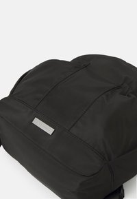Mochila de nylon negra con una superficie suave, que presenta un diseño minimalista, una placa de logo plateada y detalles de costura.