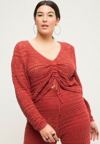 Rustfarbenes Strickoberteil mit V-Ausschnitt, langen Ärmeln und einem gerafften Frontdetail mit Kordelzug. Verfügt über ein strukturiertes Netzdesign.