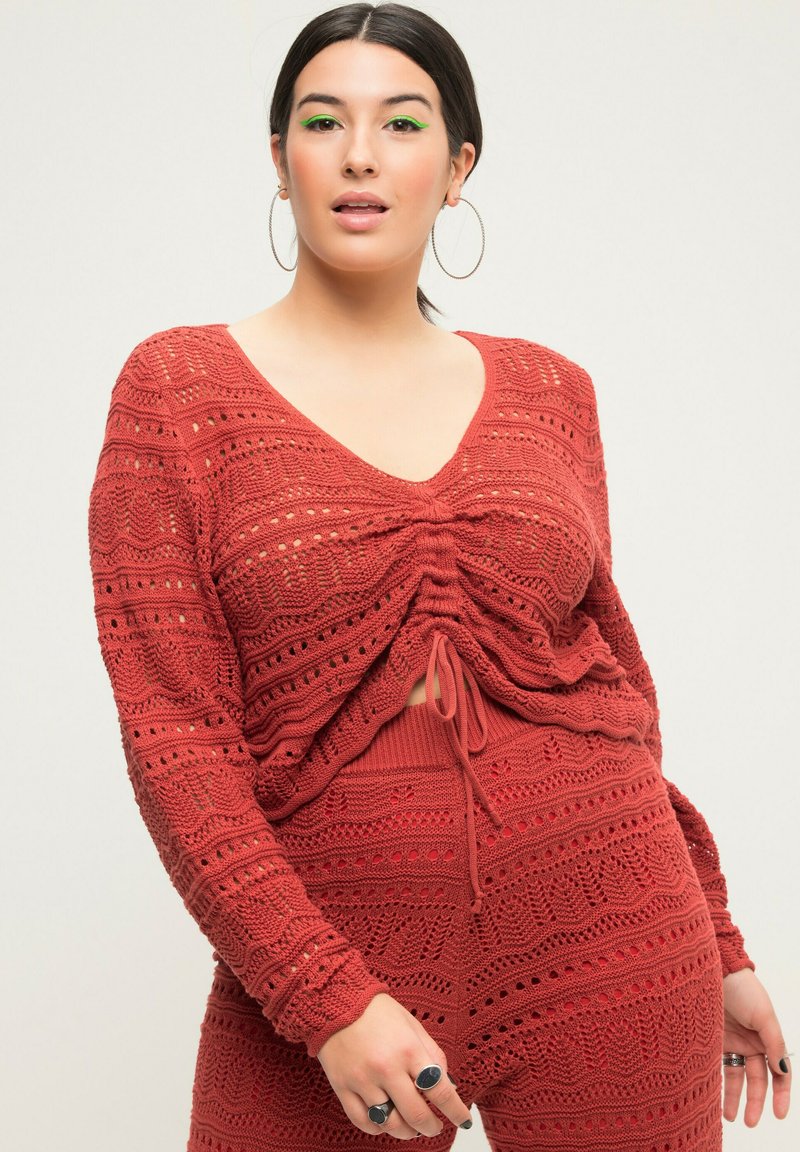 Rustfarbenes Strickoberteil mit V-Ausschnitt, langen Ärmeln und einem gerafften Frontdetail mit Kordelzug. Verfügt über ein strukturiertes Netzdesign.